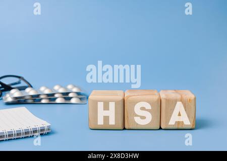 Holzblock mit Text HSA Bedeutung Gesundheit Sparkonto mit Brille, Notizbuch, Pillen hinter auf blauem Hintergrund Stockfoto