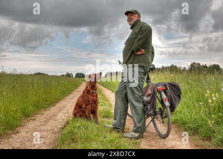 Ein Senior mit seinem Hund steht lächelnd neben seinem Fahrrad auf einem idyllischen Weg zwischen zwei Wiesen. Stockfoto