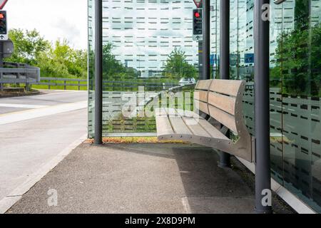 Moderne Bushaltestelle mit Holzbank am Tag an einem sonnigen Tag. Zwei verschwommene rote Lichter von Verkehrslichtern im Hintergrund. Stockfoto