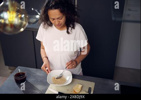 Draufsicht lockige, multiethnische Frau mittleren Alters, die zu Hause ein trockenes Frühstück zubereitete, einen Löffel in der Hand hielt und Haferflocken mit Milch mischte, am Set stehend Stockfoto
