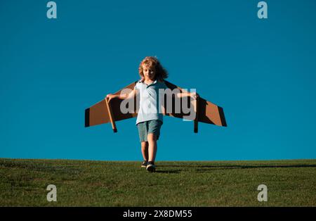 Kind, das im Flugzeug fliegt, machte Handwerk aus Pappflügeln. Traum, Phantasie, Kindheit. Reise- und Sommerurlaubskonzept. Kleiner Junge Pilot gegen einen blauen Stockfoto