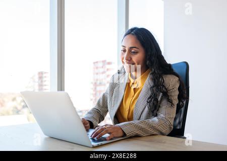 Junge lateinische Geschäftsfrau lächelt und arbeitet mit Laptop in ihrem Büro. Weibliche Führungskraft und Finanzkraft. Stockfoto