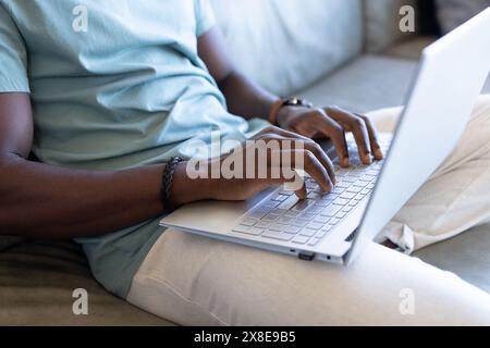 Ein afroamerikanischer Mann, der zu Hause auf dem Laptop tippt und eine Uhr trägt. Mit kurzen schwarzen Haaren und einem hellblauen T-Shirt, das sich intensiv auf das Thema konzentriert Stockfoto