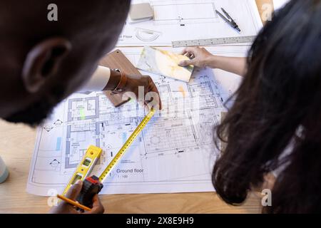 Ein vielfältiges Team arbeitet an Architekturzeichnungen am Schreibtisch mit Fokus und Zusammenarbeit. Afroamerikanischer Mann mit Maßband, während indische fema Stockfoto