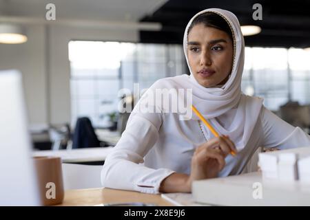 Im modernen Büro arbeitet eine indische Frau mit weißem Hijab und weißer Bluse. Bleistift halten und nachdenklich auf den Computerbildschirm schauen, umgeben von Stockfoto