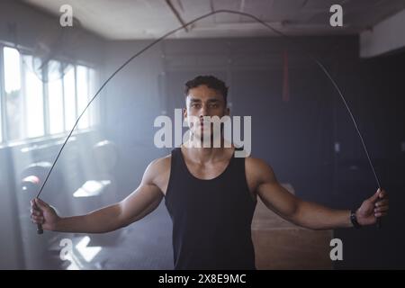 Starker und fitter birassischer Mann im Fitnessstudio, der in schwach beleuchtetem Fitnessstudio schnappt. Sonnenlicht strömt durch große Fenster und wirft Schatten auf Fitnessgeräte und Wände, u Stockfoto