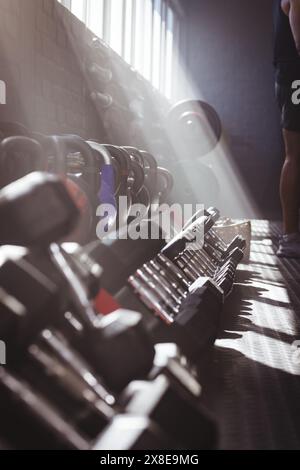 Kräftiger und fitter junger Mann, der im Fitnessstudio steht, mit verschiedenen Gewichten im Vordergrund, Kopierraum. Sonnenlicht, das durch die Fenster strömt, beleuchtet die Ausrüstung Stockfoto