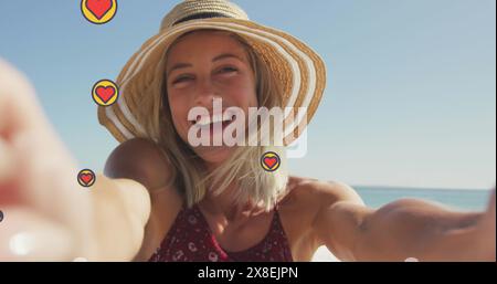 Bild von herzförmigen Ikonen, wunderschöner kaukasischer Frau, die Kamera hält und am Strand aufzeichnet Stockfoto