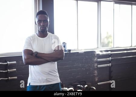 Starker und fitter afroamerikanischer Mann, der mit überkreuzten Armen im Fitnessstudio steht, Kopierraum. Sonnenlicht strömt durch große Fenster und beleuchtet Gewichtsregale Stockfoto