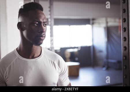 Starker und passender afroamerikanischer Mann im Fitnessstudio mit weißem Hemd, Kopierraum. In der Nähe von Fitnessgeräten mit fokussiertem Ausdruck, mit Fitnessgeräten und Stockfoto
