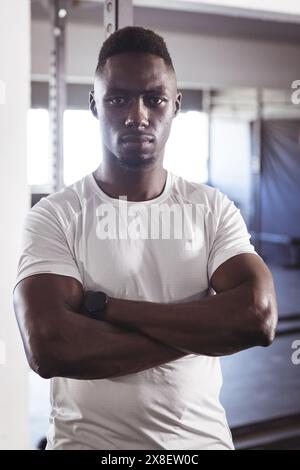 Starker und fitter afroamerikanischer Mann im Fitnessstudio, der mit überkreuzten Armen steht. Er ist in einem modernen Fitnessraum mit verschiedenen Gewichten und Geräten, was energetische atmos schafft Stockfoto