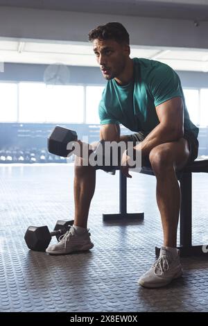 Starker und passender birassischer Mann im Fitnessstudio, der Hanteln hebt, während er auf der Bank sitzt Stockfoto