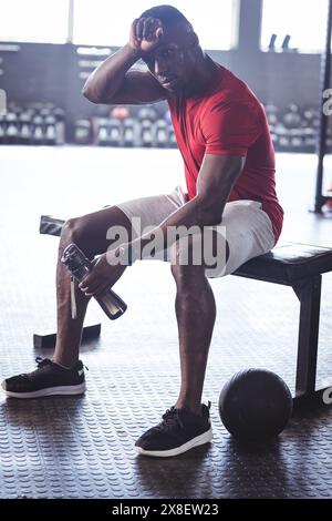 Im Fitnessstudio ruhte ein afroamerikanischer Fitnessfan Mitte 20 auf der Turnbank. Er ist in einem modernen Fitnessstudio mit verschiedenen Trainingsgeräten im Hintergrund, Stockfoto