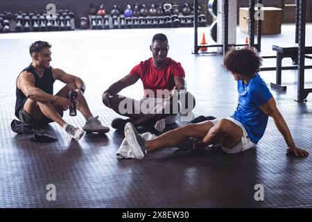 Im Fitnessstudio sitzen drei junge, fitte, vielfältige Männer auf dem Fitnessraum, plaudern und entspannen. Umgeben von Kettlebells und Gewichten in einem geräumigen, gut beleuchteten Fitnessraum, un Stockfoto