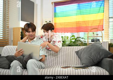 Niedliches, junges schwules Paar, das zusammen im Wohnzimmer einen Laptop benutzt, mit der LGBT-Stolz-Flagge in Regenbogenform Stockfoto