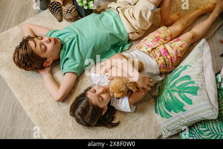 Blick von oben auf zwei Kinder mit geschlossenen Augen, die sich über dem Teppich in gemütlichem Innenraum mit warmer Atmosphäre entspannen. Stockfoto