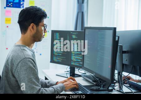 Der Prompt-Ingenieur des asiatischen Mannes entwickelt eine Codierungs-App mit Softwaredaten, die vor dem Computermonitor im Büro sitzen Stockfoto