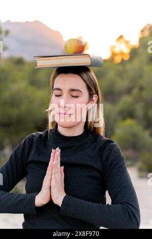 Frau, die mit einem Buch und einem Apfel auf dem Kopf meditiert. Körper und Geist im Gleichgewicht, gesunde Gewohnheiten und Wohlbefinden Stockfoto