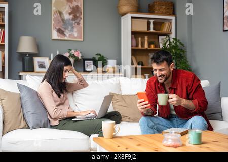 Einfaches Leben. Glückliches junges Paar, Mann und Frau sitzen entspannt auf der Couch im Wohnzimmer, lesen Buch trinken Tee zusammen, surfen im Internet, beruhigen Stockfoto