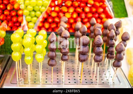 Erdbeeren in Schokoladenglasur und Trauben in Zuckerglasur auf Spießen auf dem Nachtmarkt. Street Food, leckeres Dessert. Stockfoto