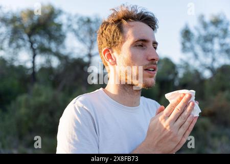 Kranker Mann umgeben von der Natur bläst sich die Nase und niest für Pollenallergiesymptome Stockfoto