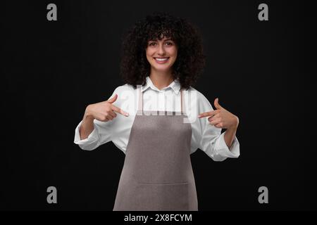 Glückliche Frau, die auf Küchenschürze auf schwarzem Hintergrund zeigt. Modell für Design Stockfoto