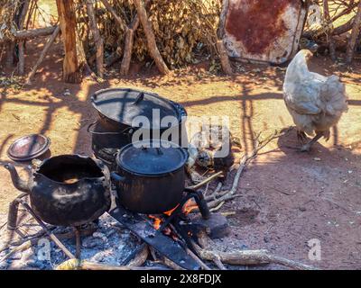 Dorf traditionelle afrikanische Außenküche, dreibeiniger gusseiserner Topf, Teekanne oben auf offenem Kaminfeuer, Huhn und Babys, die im Th herumlaufen Stockfoto