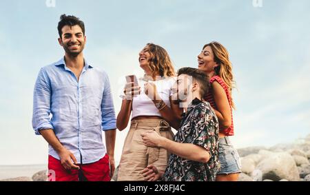 Eine Gruppe von Freunden genießt einen unterhaltsamen Moment am Strand, lacht und verbindet sich über ein Smartphone. Das Wesen des Sommers, der Freundschaft und der Freude darin einfangen Stockfoto