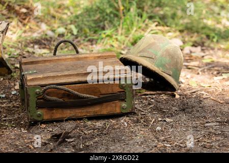 Nahaufnahme. Alte deutsche Militärmunitionsbox aus Holz auf dem Boden. Im Hintergrund ein Wehrmachtshelm m35 in Tarnhülle Stockfoto