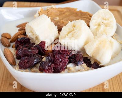 Nahaufnahme einer nahrhaften Haferflockenschale mit Erdnussbutter, Bananen und Preiselbeeren, serviert in einer weißen Schüssel auf einem Holzbrett. Stockfoto