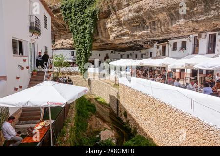 Restaurants und Cafés drängen sich unter einem Felsüberhang entlang der Terrazas Calle Cuevas del Sol in den einzigartigen Pueblos blanco von Setenil de las Bodegas, Spanien. Die Bewohner des kleinen Dorfes Setenil leben seit dem neolithikum in Höhlenhäusern. Stockfoto