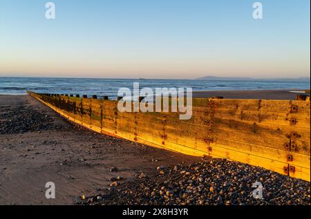 Hölzerner Wellenbrecher, der sich bei Sonnenuntergang an einem Kiesstrand entlang erstreckt, mit einem ruhigen Meer und fernen Bergen unter klarem Himmel Stockfoto