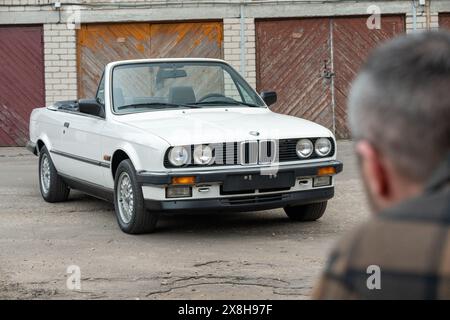 Riga, Lettland - 1. April 2024: Ein Mann, der auf sein weißes klassisches Cabriolet BMW E30 mit geöffnetem Dach blickt. Alte Garagen im Hintergrund. Stockfoto