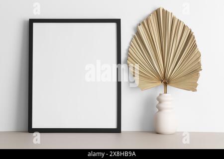 Schwarzes Rahmenmockup mit Palmblattdekoration auf dem beigefarbenen Tisch. Stockfoto