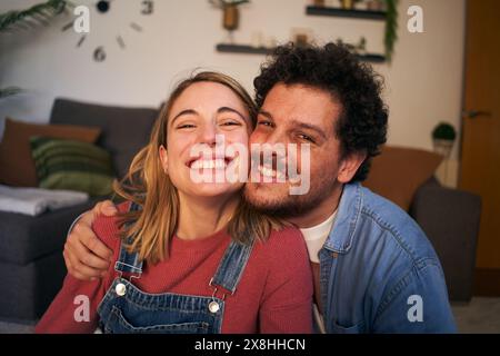 Portrait zwei fröhliche, verliebte junge Menschen, die im Wohnzimmer auf die lächelnde Kamera schauen. Weißes glückliches Paar Stockfoto