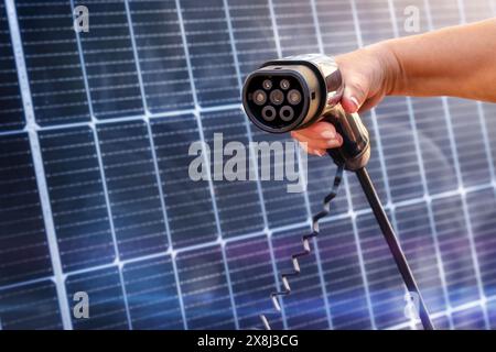 Die Hand einer Frau hält ein EV-Ladegerät für ein Auto in der Nähe von Solarpaneelen. Alternative Energie Stockfoto