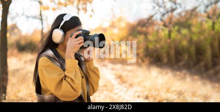 Junge schöne Fotografin in Kopfhörern spaziert in lässiger Kleidung durch den Herbstwald. Hält eine professionelle Kamera in den Händen, nimmt Stockfoto