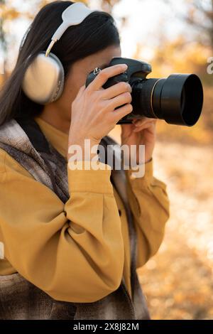 Junge schöne Fotografin in Kopfhörern spaziert durch den Herbstwald in lässiger Kleidung. Hält eine professionelle Kamera in den Händen, macht Fotos Stockfoto