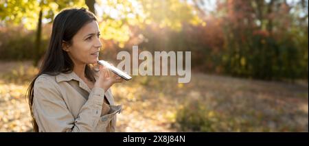 Hispanische Frau mit Sprachassistenten auf dem Handy, die mit dem Smartphone im Herbstwald spricht Stockfoto