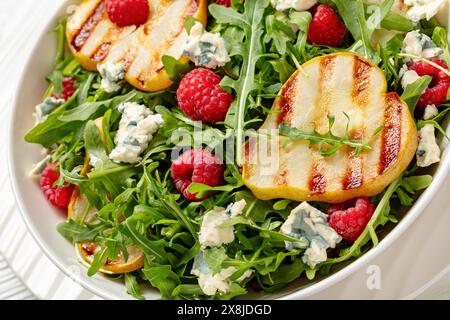 Nahaufnahme gegrillter Birnensalat mit Blauschimmelkäse, Rucola und frischen Reifen Himbeeren auf weißem Teller auf weißem Holztisch Stockfoto
