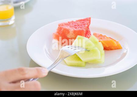 Eine Nahaufnahme eines Tellers mit Scheiben Wassermelone, Papaya und Melone. Eine Hand, die eine Gabel hält, nimmt ein Stück Melone auf. Es gibt ein Glas orange j Stockfoto