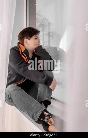 Traurige junge Frau sitzt auf der Fensterbank und bewundert die Winterlandschaft. Die Frau ist in ihren Gedanken verloren. Das Konzept der Einsamkeit Stockfoto