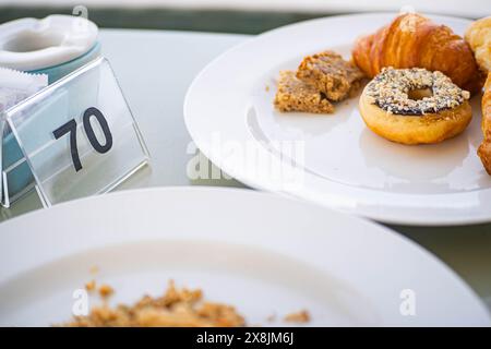Nahaufnahme eines Tisches mit einem Teller mit Gebäck einschließlich Croissant, Donut mit Nüssen und einem Stück Kuchen. Die Tabellennummer 70 ist im Backgr sichtbar Stockfoto