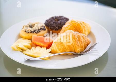 Ein weißer Teller mit einer Auswahl an Frühstücksgebäck und Obst, darunter ein Croissant, zwei Donuts, Ananasscheiben und ein Stück Papaya mit einer Gabel Stockfoto
