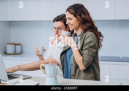 Junge Paare stehen in ihrer modernen Küche, halten Kaffeetassen und lächeln, während sie auf den Laptop schauen. Der Mann trägt eine Brille und hat Kopfhörer um den Hals, während die Frau neben ihm steht und eine entspannte und fröhliche Atmosphäre zeigt. Stockfoto