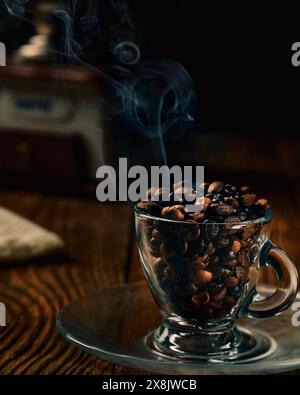 Kaffeebohnen unterschiedlicher Röstgrade in einer transparenten Kaffeetasse auf einem Holztisch mit manueller Kaffeemühle und Rauch im Hintergrund Stockfoto