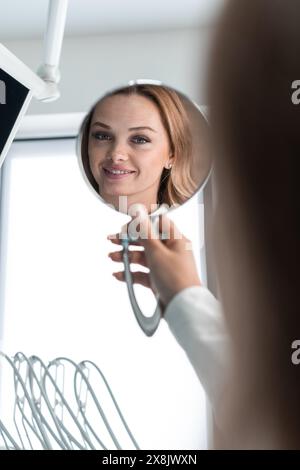 Die Patientin posiert mit einem Spiegel in der Hand und zeigt ihr Selbstvertrauen und ihre Freude an dem makellosen Lächeln, das ihr der Zahnarzt schenkte. Ein Lächeln Stockfoto