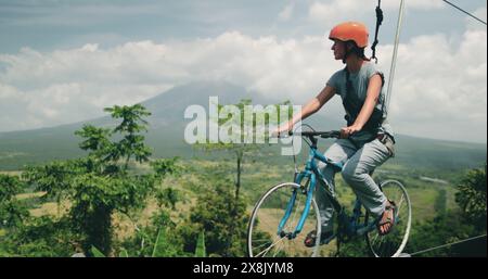 Eine Womani, die auf einem Fahrrad auf einer Seilrutsche über die wunderschönen Vulkankrater-Seen auf den Philippinen fährt. Die waghalsige Handlung kombiniert Radfahren und Seilrutschen für ein abenteuerliches Erlebnis. Stockfoto