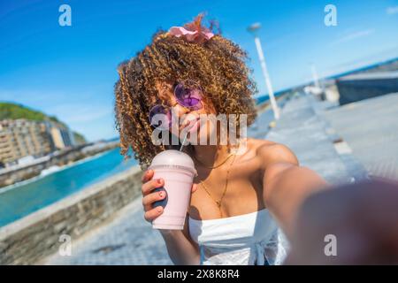 Persönliche Perspektive einer coolen jungen lateinischen Frau mit lockigem Haar und Sonnenbrille, die Milchshake trinkt, beim Selfie auf der Promenade Stockfoto