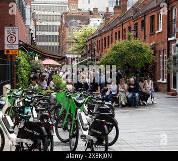 Überfüllte Sitzplätze im Freien in einer gemütlichen urbanen Gasse mit E-Bikes im Vordergrund und Gebäuden im Hintergrund. Fußgänger- und Radfahrbereichsschild. Stockfoto